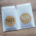Two small glassine bags with personalised kraft monogram stickers on a wooden surface