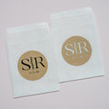 Two white glassine bags with 'SR' initials and a date printed on kraft paper labels against a light pink background.