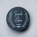 Round black sticker with white text 'Sarah & Adam 31.07.25' in a leaf frame on a light grey background