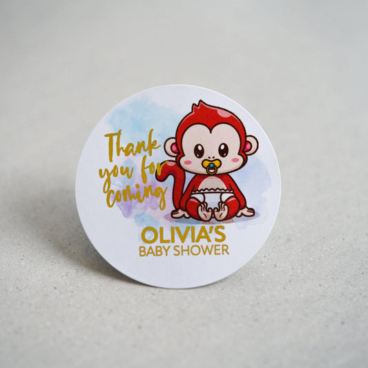 A baby shower sticker with a lovely colour print of a monkey sitting on the  floor. Thank you for coming written in a handwriting font next to the monkey and the mother to be name on the bottom. The writing is finished with metallic foil