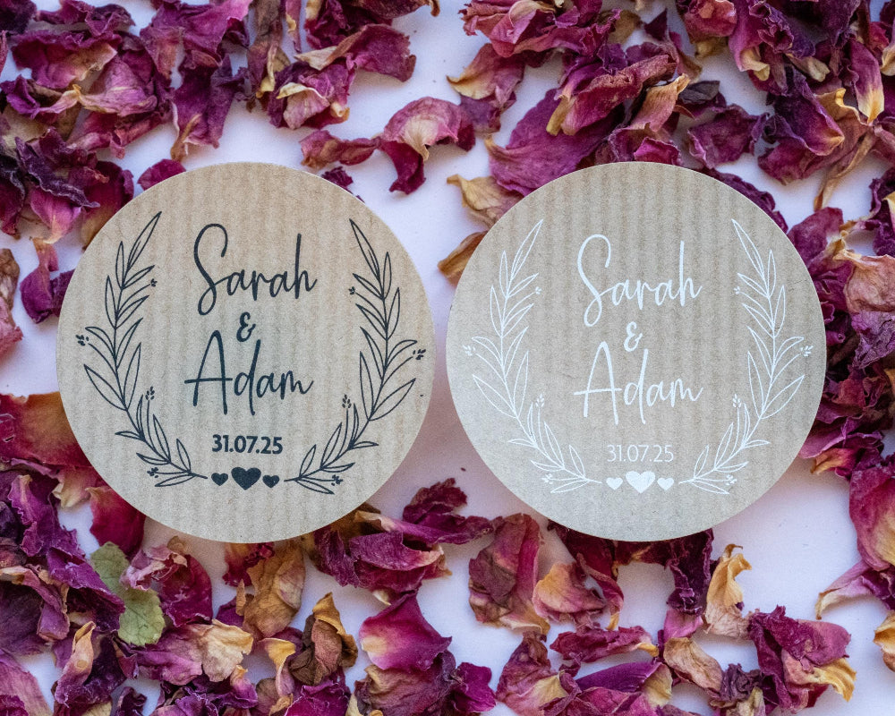Two white glassine envelopes with custom kraft wedding stickers featuring leaf patterns and personalised text 'Sara & Adam'. They are placed on a pink background with scattered rose petals.