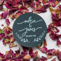 Round black sticker with 'Maria & James' and date printed using white ink surrounded by dried pink and red flowers