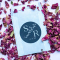 A round sticker with a monogram print, placed on a surface scattered with purple rose petals. The sticker features text and a leaf design element.