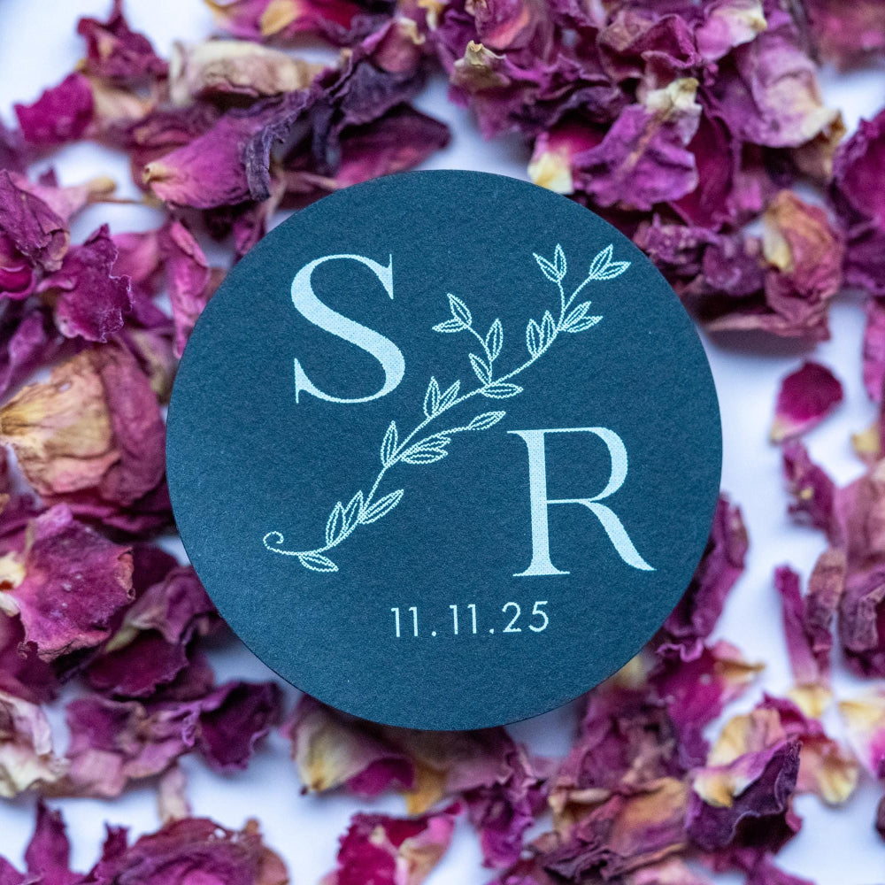 A round sticker with a monogram print, placed on a surface scattered with purple rose petals. The sticker features text and a leaf design element.