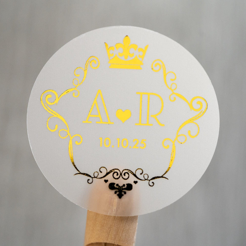 A round clear sticker with a crown frame monogram design, featuring gold print on a white background.