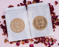 Two white glassine envelopes with custom kraft wedding stickers featuring leaf patterns and personalised text 'Sara & Adam'. They are placed on a pink background with scattered rose petals.