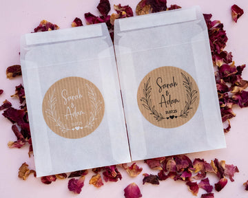 Two white glassine envelopes with custom kraft wedding stickers featuring leaf patterns and personalised text 'Sara & Adam'. They are placed on a pink background with scattered rose petals.