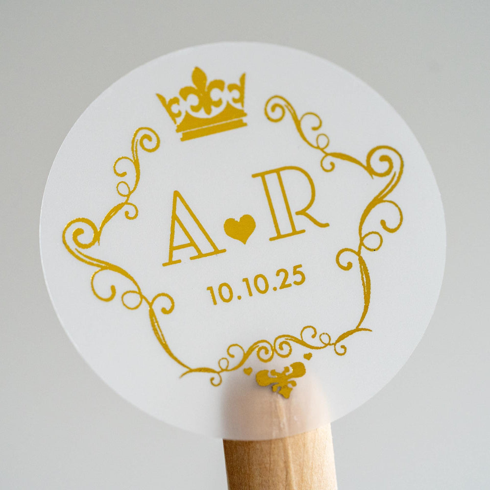 A round clear sticker with a crown frame monogram design, featuring gold print on a white background.